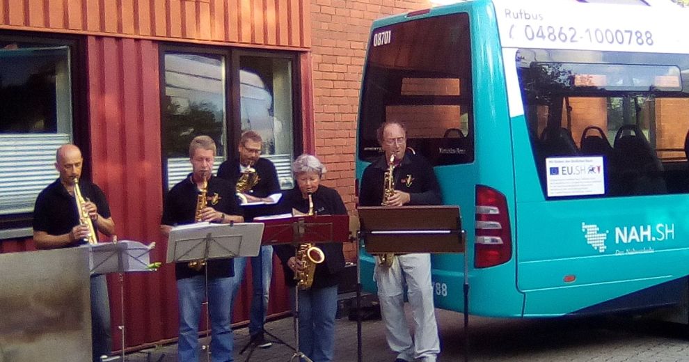 Fünf Personen stehen Saxophon spielend neben einem Kleinbus. 