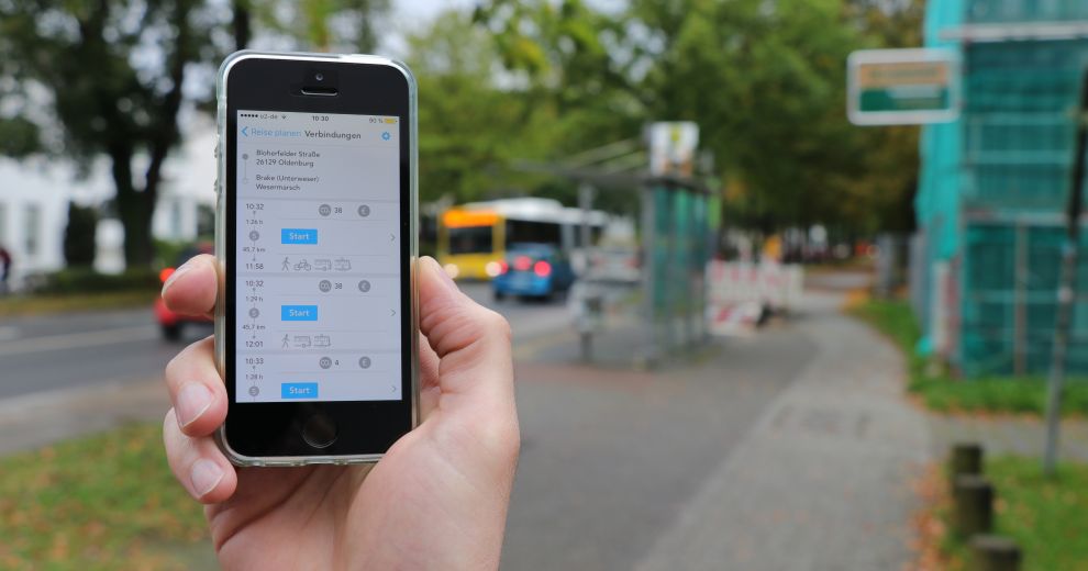 Kunde hält vor einer Bushaltstelle sein Mobililtelefon mit der geöffneten App in den Händen