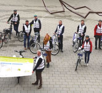 Eine Gruppe von Personen in passenden Westen und mit Fahrrädern hält ein Banner hoch.