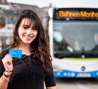 Eine junge Frau, die vor einem Bus steht und den Monheimpass in die Kamera hält.